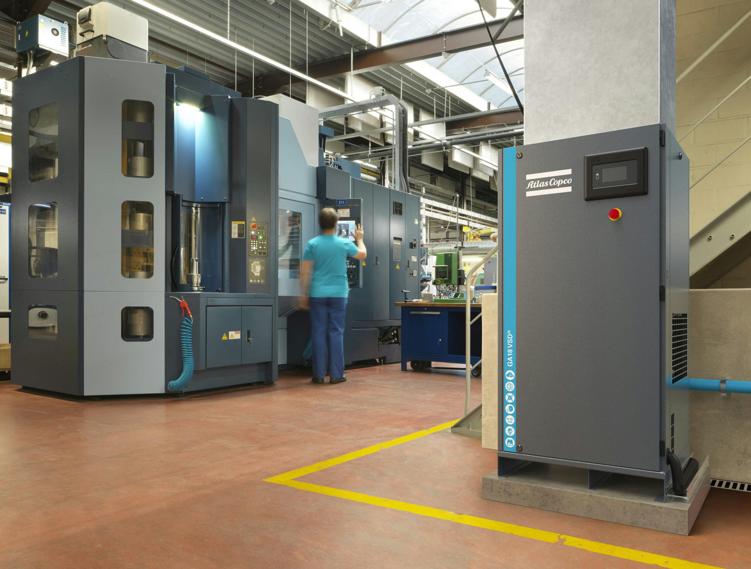 Kompressor som er plassert i produksjonshall, medarbeider jobber med produksjon i CNC maskin. Kompressor er koblet til blå air-net rør.